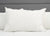 A Down Etc. 25% White Goose Down / 75% White Goose Feather Pillow sits upright against a light gray upholstered headboard, centered on a neatly made bed with a matching white comforter. A small fabric tag is visible on the pillow’s lower right corner.