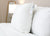 Two Keeco Best Western® Everest Pillows | Soft Polyester Pillows, each with tags, rest against a white bedspread on a tidy bed. A softly lit lamp sits on a wooden nightstand, set against a beige wall for a calm, minimal bedroom look.