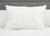 A Pillowtex White Goose Down & Feather Pillow (75% feather/25% down) sits upright on a white quilted comforter, framed by a gray headboard. The soft, plush pillow has slightly wrinkled white fabric with no visible patterns.