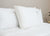 The Keeco Best Western® Aurora Medium Polyester Pillow rests on crisp white bedding, with a light wall and a nightstand plant in the background, creating a calm and inviting atmosphere. Hypoallergenic and perfect for a restful night.