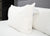 A Pillowtex Triple Core Lyocell Pillow | Medium Support, with brand and care tags visible, rests against a gray upholstered headboard on white sheets, offering excellent neck support. Another pillow lies flat behind it amid dark furniture.