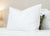 A Down Dreams Classic Soft Pillow by Manchester Mills, featured at many hotels, in white with a tag is on a white bed. Behind it is a smaller pillow with a green floral pattern. The softly lit background is a plain beige wall.