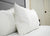 Two Down Etc. 25% White Goose Down / 75% Feather Pillows with smooth cases rest against a gray upholstered headboard on a neatly made bed with white sheets. A closed white door with a black handle is visible in the bright background.