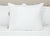 A Pillowtex Premium Polyester Pillow | Extra Firm from Pillowtex sits upright on a neatly made bed with a white quilted comforter. The overstuffed pillow has a care tag on its right side. The scene is well-lit and minimalistic.