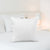 A pristine Pillowtex Pillow Insert filled with White Duck Feather & Down stands against a backdrop of crisp bedding in a brightly lit, minimalist bedroom, exuding a tranquil and inviting ambience for rest.