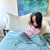A woman in pink floral pajamas sits on an unmade bed with a blue quilt, smiling at the Hollander Holiday Inn® Soft and Firm Polyester Pillow Combo Pack (2 pillows). A nightstand, white lamp, and abstract teal and blue painting add to the cozy scene.