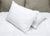 Two Green Label Soft Pillows by Keeco, featured at Many Choice® Hotel brands, rest on a white quilted bed. Taupe pillows add contrast behind. The front soft support pillow stands upright, ideal for stomach and back sleepers, with a green tag visible.
