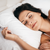 A dark-haired woman in a white tank top lies peacefully on a bed, smiling with her eyes closed as she rests her head and hand on the Keeco Holiday Inn® Infinity Pillow | Extra-Firm Support, creating a calm, relaxed atmosphere.