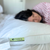 A person with dark hair lies on a bed, face slightly out of focus, resting on the Keeco Green Label Soft Pillow—featured at many Choice® Hotel Family of Brands. The white pillow shows its green and black tag; the person wears a red and white patterned top.