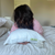 A person with wavy brown hair in a white shirt with red dots lies face-down on a white bed, hugging a Keeco Green Label Soft Pillow, featured at many Choice® Hotel brands. Another white pillow and a neutral wall with door are in the background.